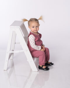 Cute toddler wearing pink overalls ad sitting on a white ladder photographed on a white background.