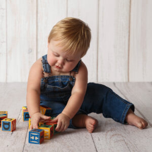 Baby Milestone Portrait of a Toddler wearing overalls and sitting on the floor with blocks.