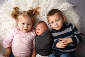 Three siblings laying on a rug including a little girl in a pink shirt and pig tails, a big brother in a strips shirt and the newborn baby