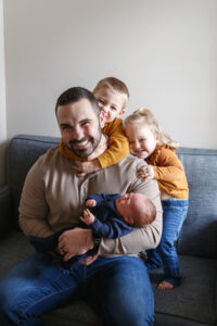 Dad sitting on a couch holding his newborn baby while older siblings cuddle behind him playfully