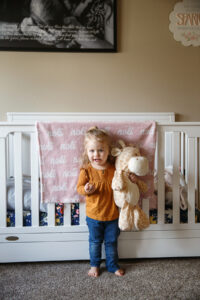 Big sister stands by the new baby's crib holding a stuffed animal