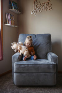 Big sister sitting on a gray chair and holding a stuffed giraffe