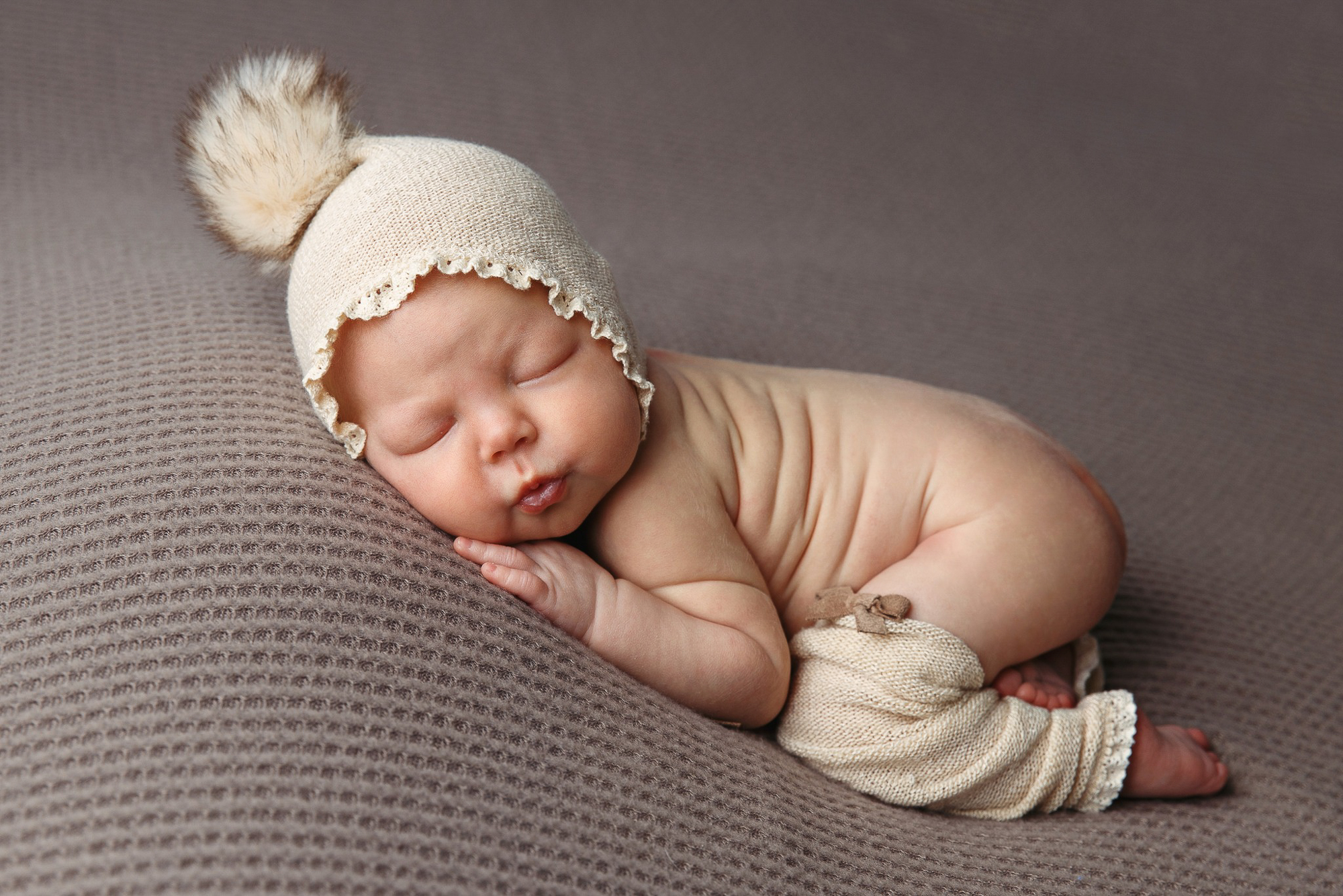 Photoshoot of a newborn laying on it's tummy with it's bottom up and wearing a beige bonnet and leggings