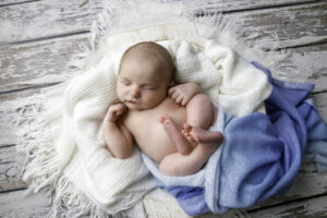 Newborn baby boy asleep on his back with his feet crossed on a white blanket with a blue blanket 