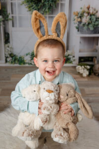 Cute little boy wearing bunny ears and holding three stuffed bunnies in front of an Easter themed background