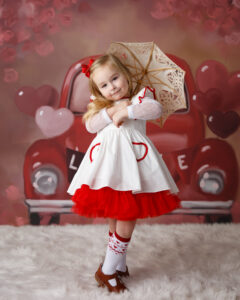 Photo of a little girl with an umbrella standing in front of a valentine themed background with a red Volkswagon car