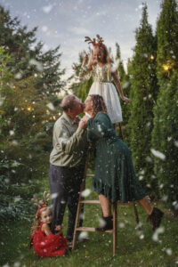 Family photo at a Christmas tree lot standing by a ladder with one child holding mistle toe over parents heads as they kiss