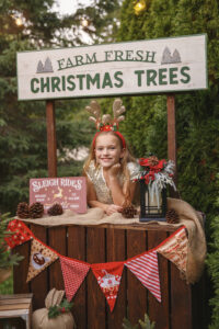 Little girl wearing antlers behind leaning on the counter of a Christmas Tree stand