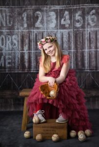 Girl wearing a fancy red dress and flower crown photographed with tennis shoes and catchers mitt on a baseball dugout background