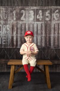 Little two year old boy wearing a baseball uniform with red hat and red socks and sitting on a bench in front of a dugout backdrop