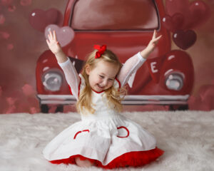 Image of a three year old girl in a cute dress sitting in front of a background with a Volkswagon Beatle car and holding her hands up with peace signs 