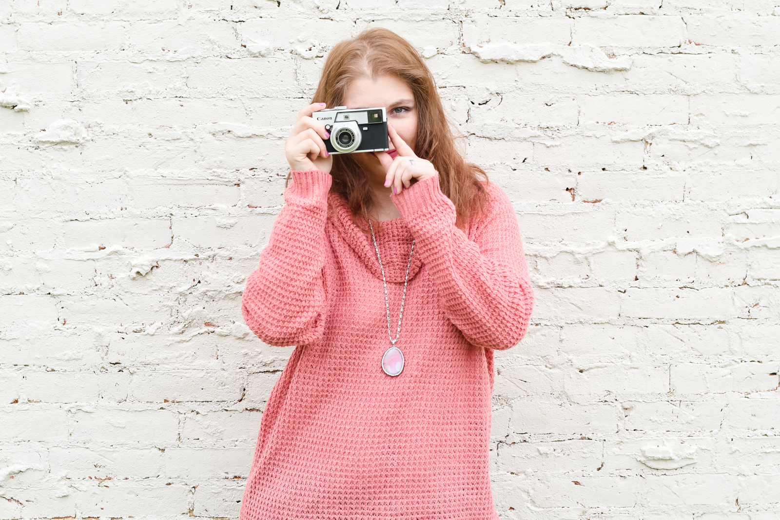 Image of a photographer in a pink sweater standing in front of a white brick wall and holding a camera.