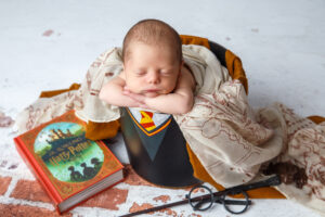 Portrait of a Newborn baby sitting in a bucket with a Harry Potter book, glasses and wand,