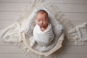 Newborn Baby wrapped in a white blanket laying in a white bowl.
