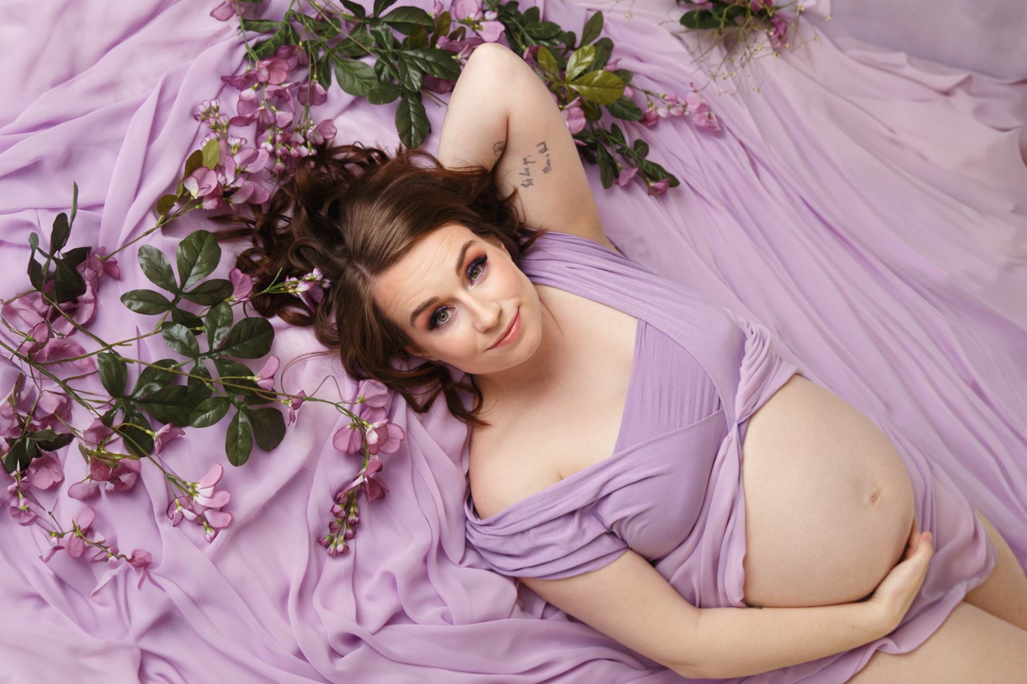 Photo of a pregnant woman laying in lavender colored fabric with purple flowers.