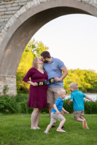 Family of four portrait. Mom and dad kiss as their young boys run around them. They are holding a sonogram of the baby who will be coming soon.