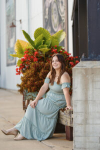 Portrait of a high school senior girl in a blue dress sitting near a large plant in downtown Beloit.