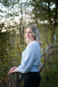 Portrait of a high school senior girl in a blue sweater standing near a split rail fence at headgate park near Janesville.