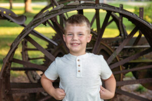 Smiling boy in a white shirt standing in front of an antique iron tractor wheel.