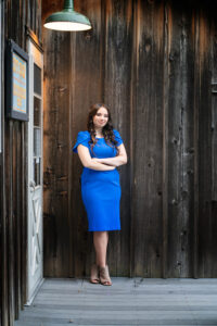 Senior Portrait of a pretty teen girl wearing a blue dress and standing against a rustic building.