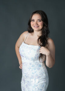 Three quarter image of a high school senior wearing a white dress and posing on a dark gray background.