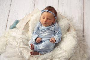 Newborn baby girl wearing a blue onesie and laying in a basket with a white rug.