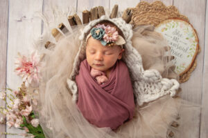 Newborn in a pink wrap laying in a basket and wearing a flowered headband during a newborn session.