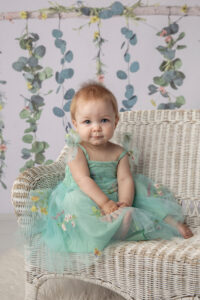 Cute one year old girl in a pretty green dress sitting on a white whicker chair.