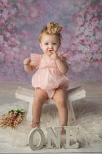 One year old girl dressed in a pink jumper sitting on a little white bench with a crown on her head.