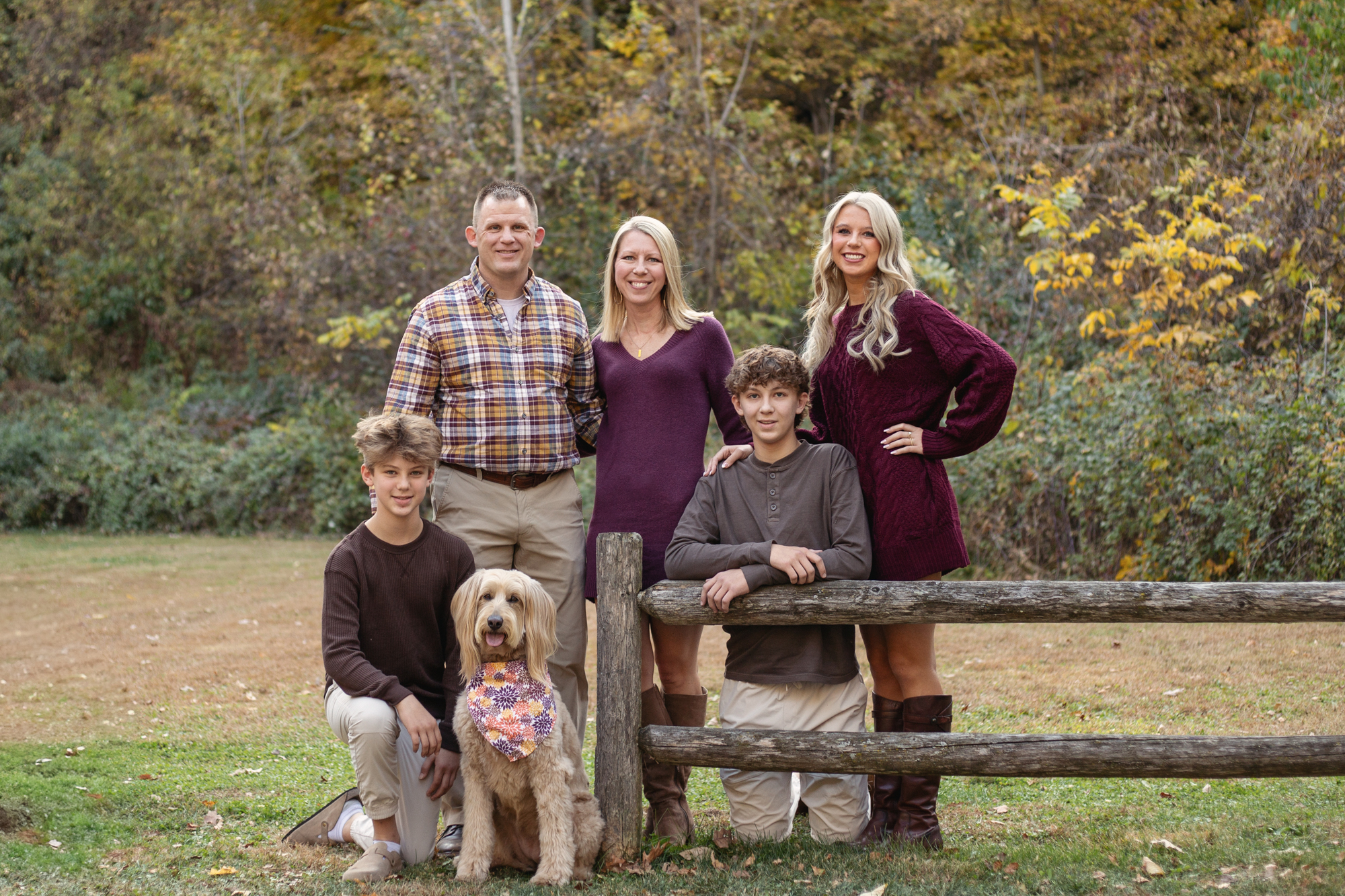 Family photo of five with older children and dog taken outdoors near a split rail fence.