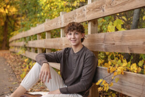 Photo of a highschool aged boy wearing a gray sweater and sitting outdoors next to a wooden fence. 