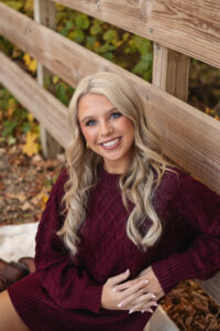 Portrait of a young woman with blond hair sitting next to a wooden fence and smiling. 