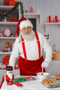 Santa wearing white shirt and suspenders standing near a table with mixing bowls like he's baking.