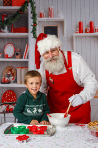 A boy in a green sweater standing with Santa who is wearing a red apron and mixing flour in a bowl like he's baking something.