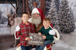 Two brothers sitting with Santa and holding a Merry Christmas sign.