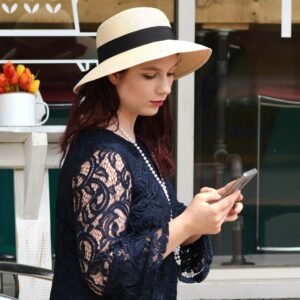 Pretty young woman wearing a navy lace dress and hat sitting outside of a coffee shot on her cell phone.