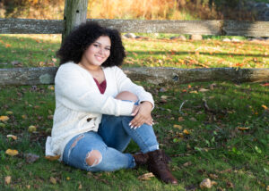 Pretty girl in a white sweater posed in front of a split rail park in the fall before her high school graduation.