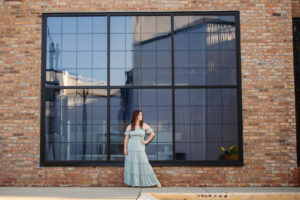 Beautiful girls in a long dress posing in front of a large window downtown for portraits taken a by professional senior photographer.