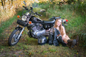 High school senior in a cute dress and leather jacket on the grass in front of her motorcycle and posing for senior portraits.