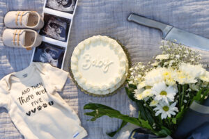 Photo of a white gender reveal cake along with a sonogram and a bouquet of white flowers sitting on a picnic blanket. 