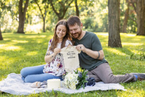 Young couple sitting on a blanket holding up a baby onesie for an image reveal session at Magnolia Bluffs in Evansville.